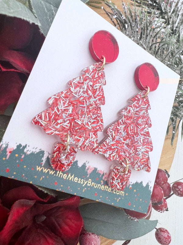 Red and white Christmas tree earrings on a branded card with a festive background.