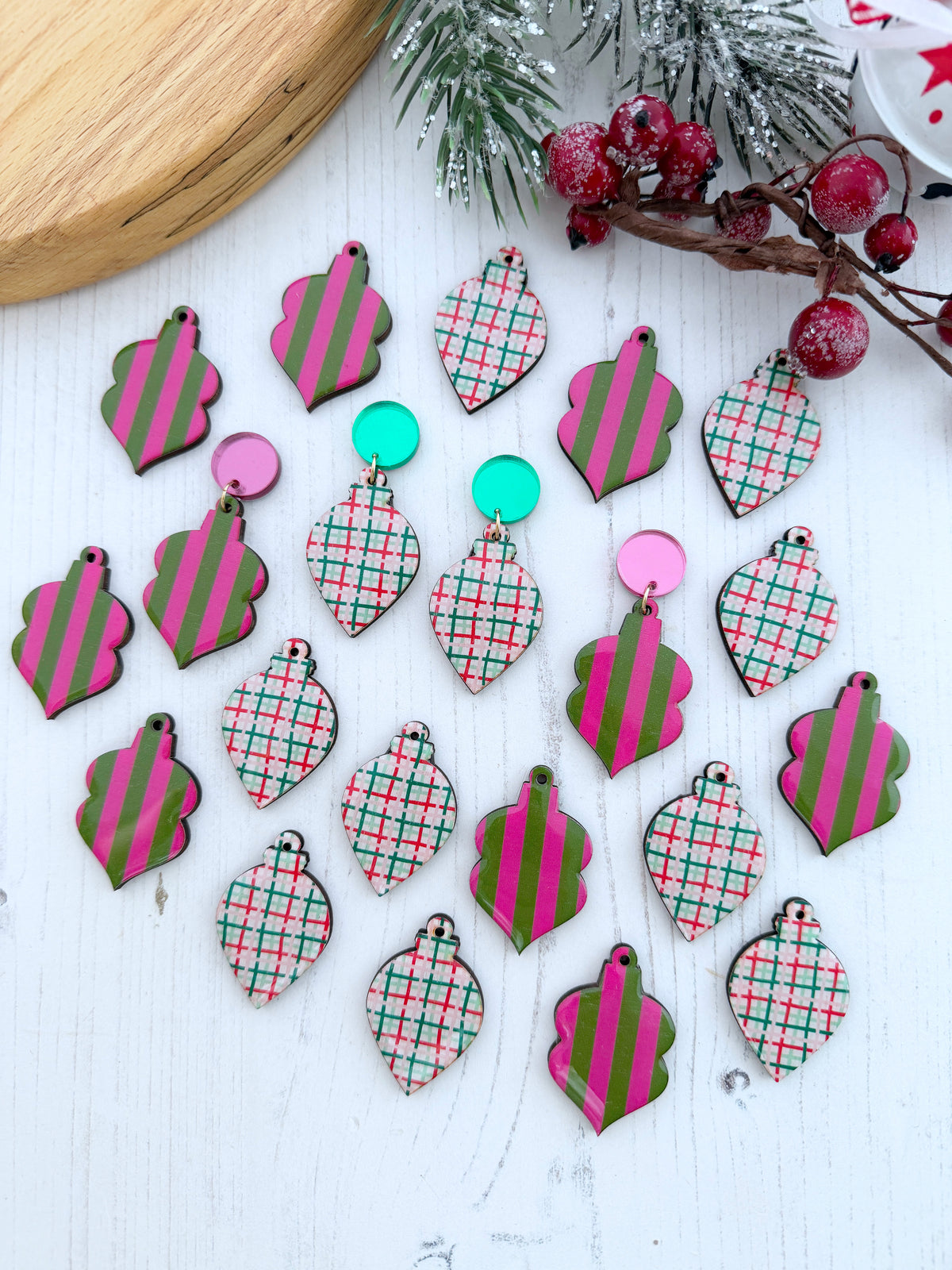 A flat lay of assorted Christmas ornament-shaped wooden earrings on a white wooden surface, featuring pink and green striped and plaid patterns, with holiday greenery and red berries as props.