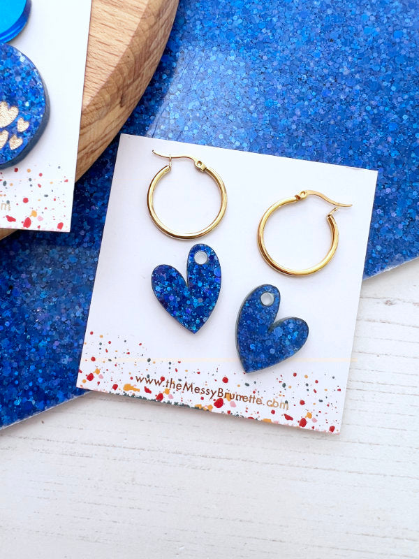 Blue heart-shaped earrings on a branded card with gold hoop earrings above, against a blue textured background.