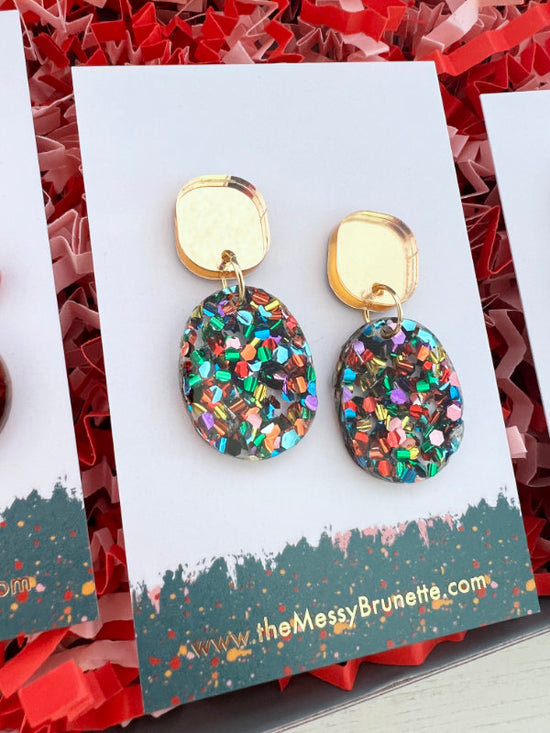 Multicolored glittery earrings on a 'the Messy Brunette' branded card with red and white paper background.
