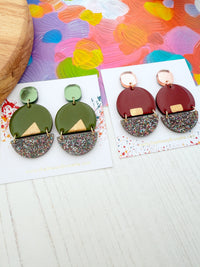 Two pairs of statement earrings displayed on white cards on a light wood surface. One pair features olive green and dark green geometric shapes with glittery multicolor lower halves and a small gold triangular accent. One other one is burgundy colour with multicoloured glitter