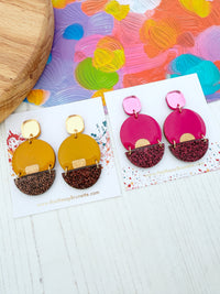 Two pairs of statement earrings displayed on white cards on a white wood surface. One pair is mustard yellow with glitter and the other is pink with burgundy pink glitter