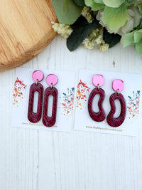 A pair of statement earrings displayed on white cards on a white wood surface, with a wooden board and green flowers as styled props. The earrings have bright pink circular studs and elongated dark pinkish-red wavy drop pieces.