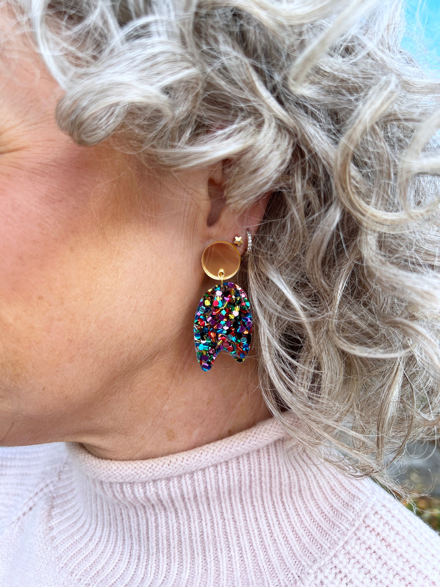 Close-up of a person wearing colourful glitter earrings with a light background