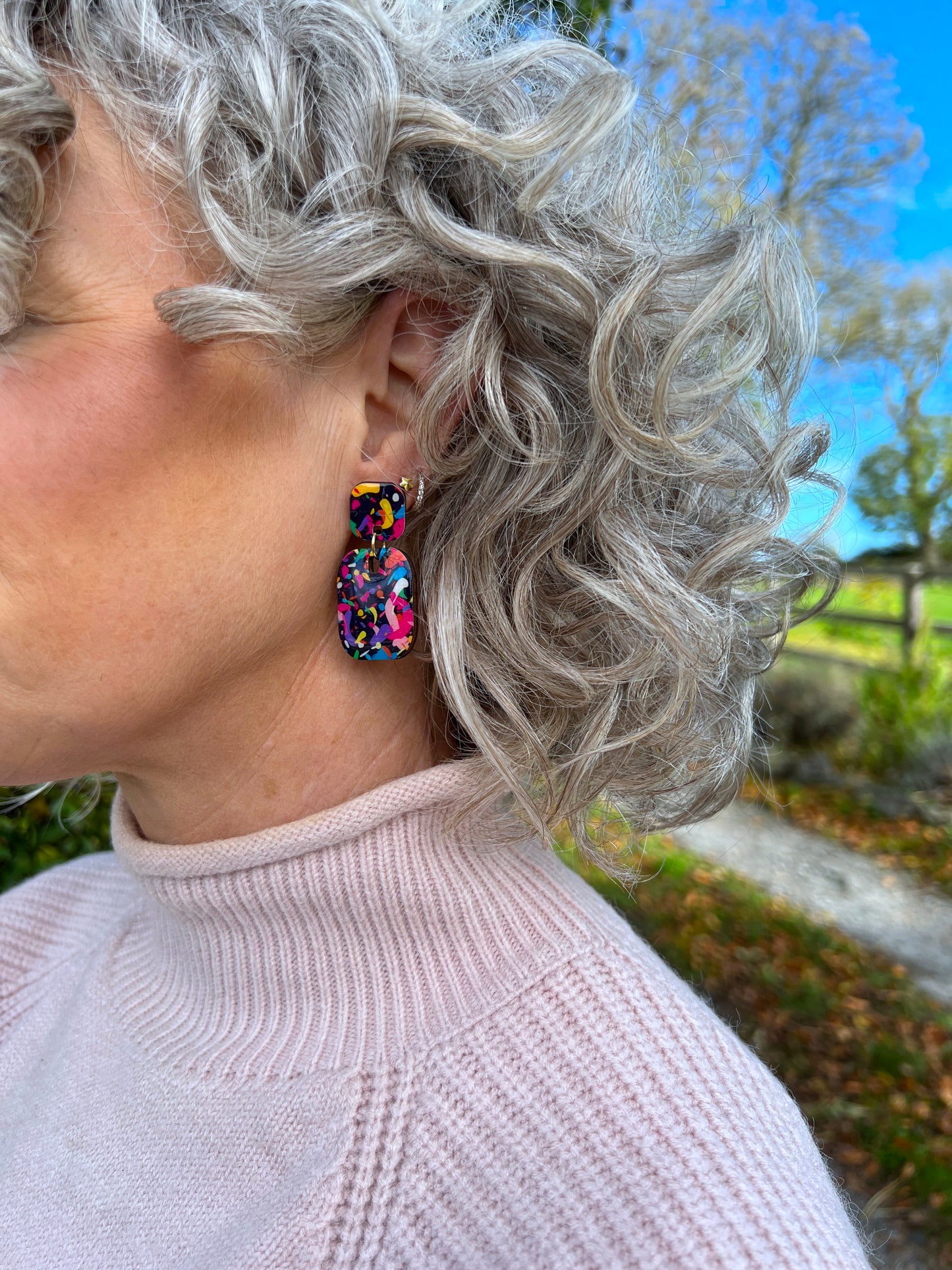 Person with curly gray hair wearing colourful wood earrings outdoors