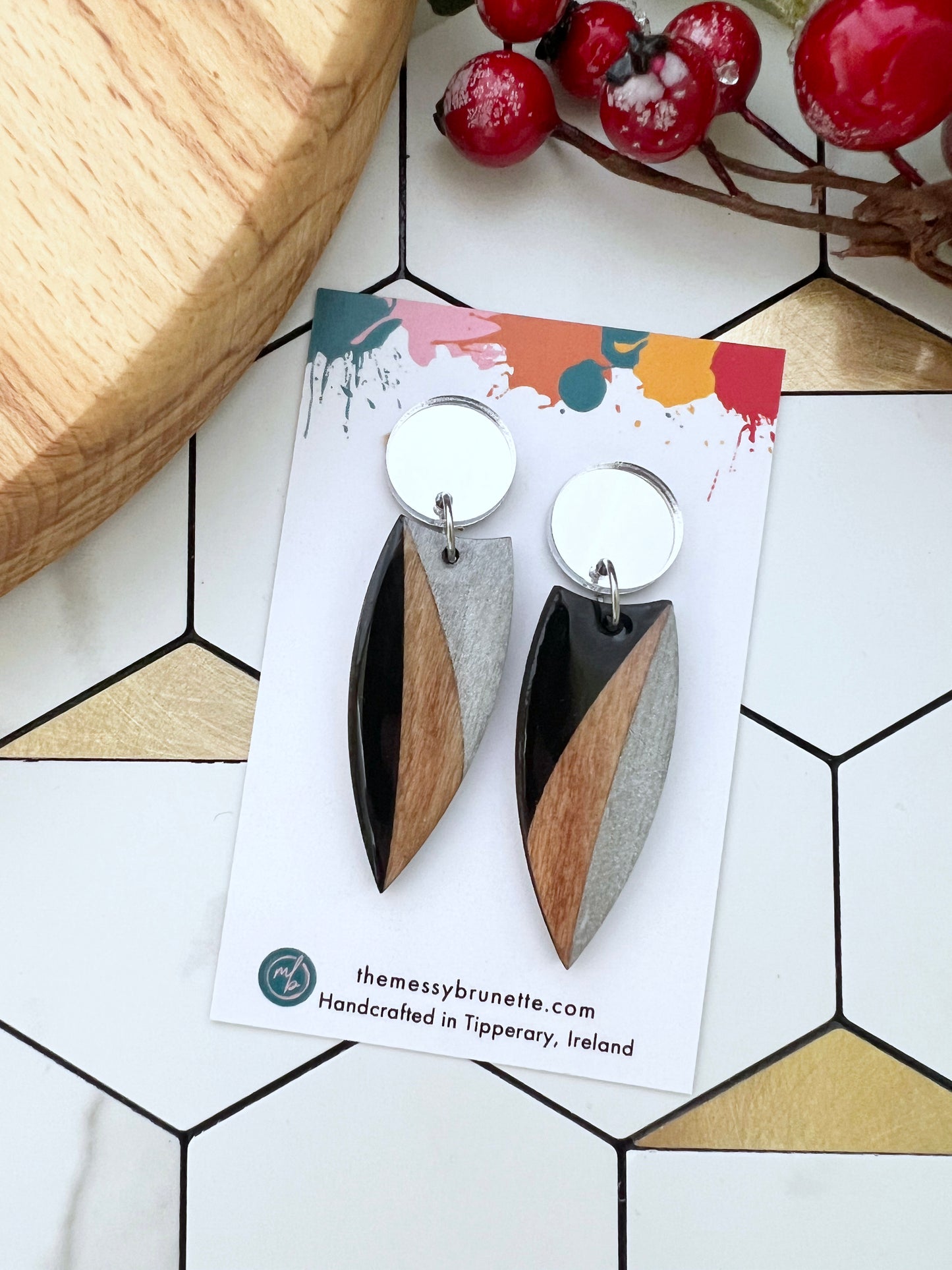 Wood earrings painted in black and silver on a colourful card with 'themessybrunette' branding, placed on a geometric patterned surface.