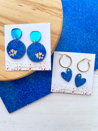 Glittery Blue and gold earrings on branded cards with a blue textured background