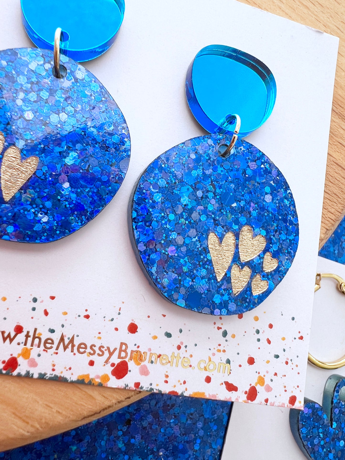 Round blue glittery earrings with gold hearts inked to the front on a branded card
