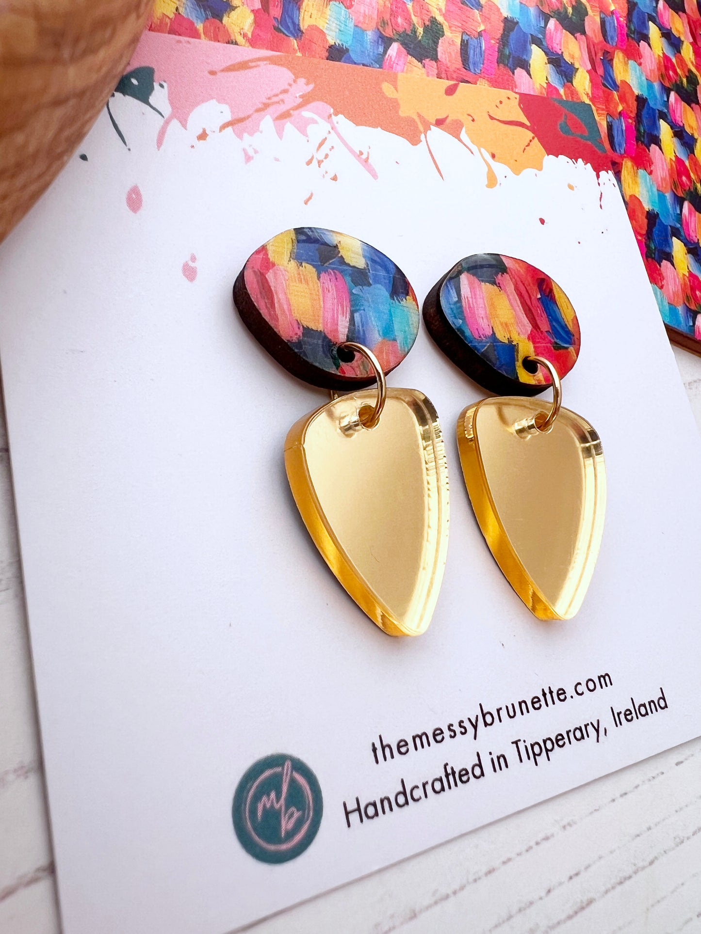 Gold and colourful earrings on a branded card with a colourful abstract design 