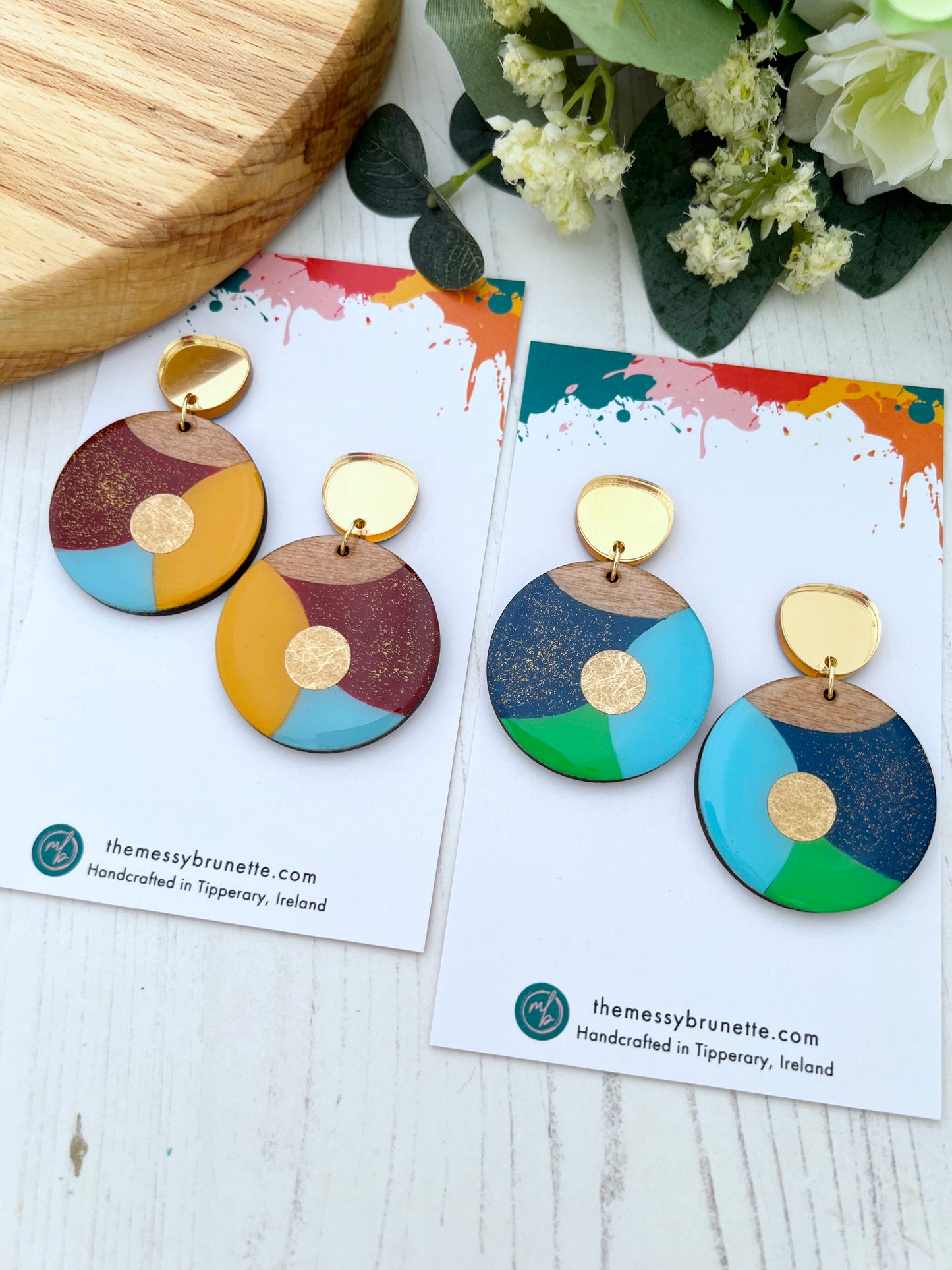 Colourful earrings on cards with 'themessybrunette.com' branding, placed on a wooden surface with flowers.