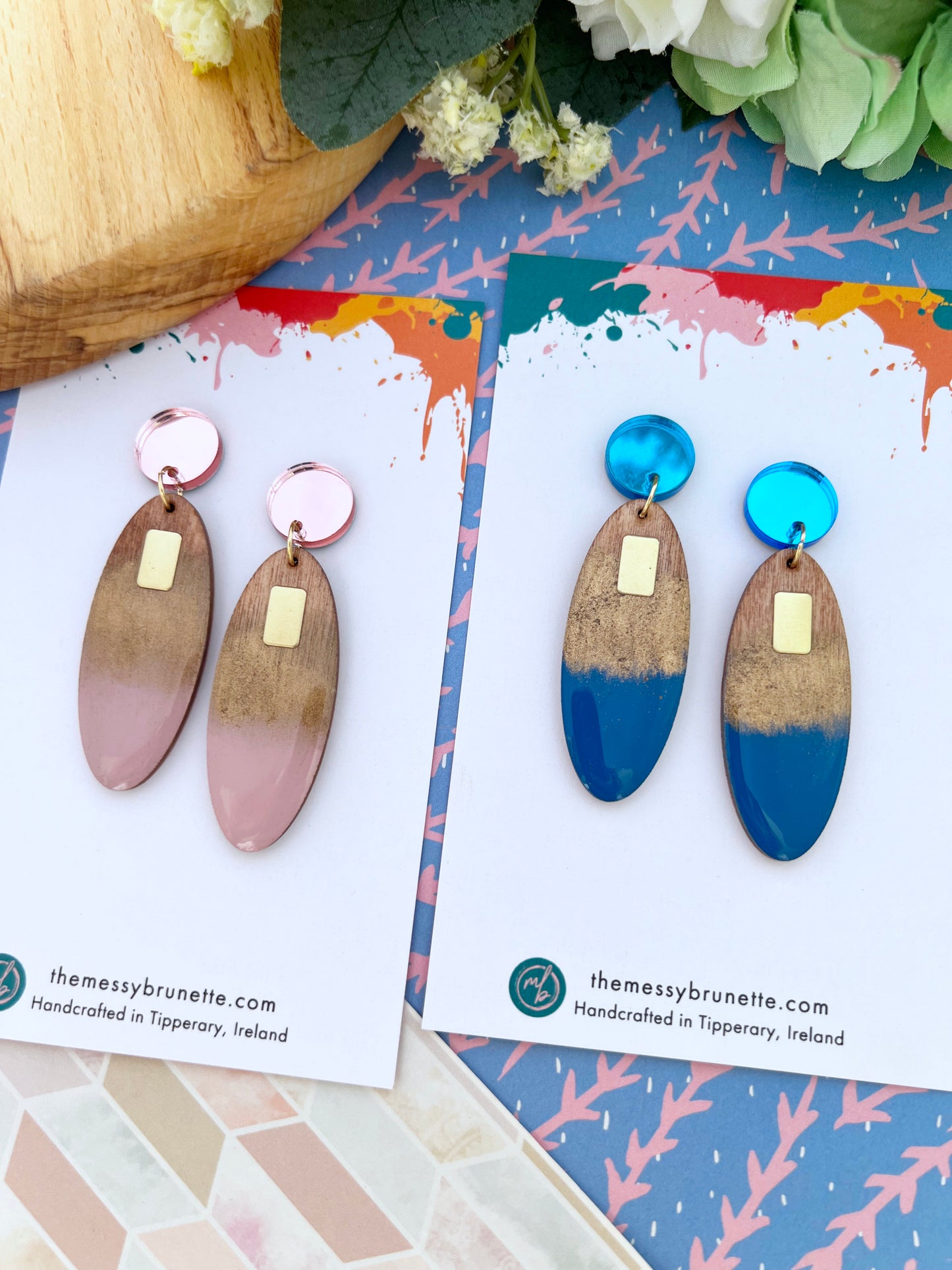 Two pairs of long oval painted wood earrings on cards with a colourful background