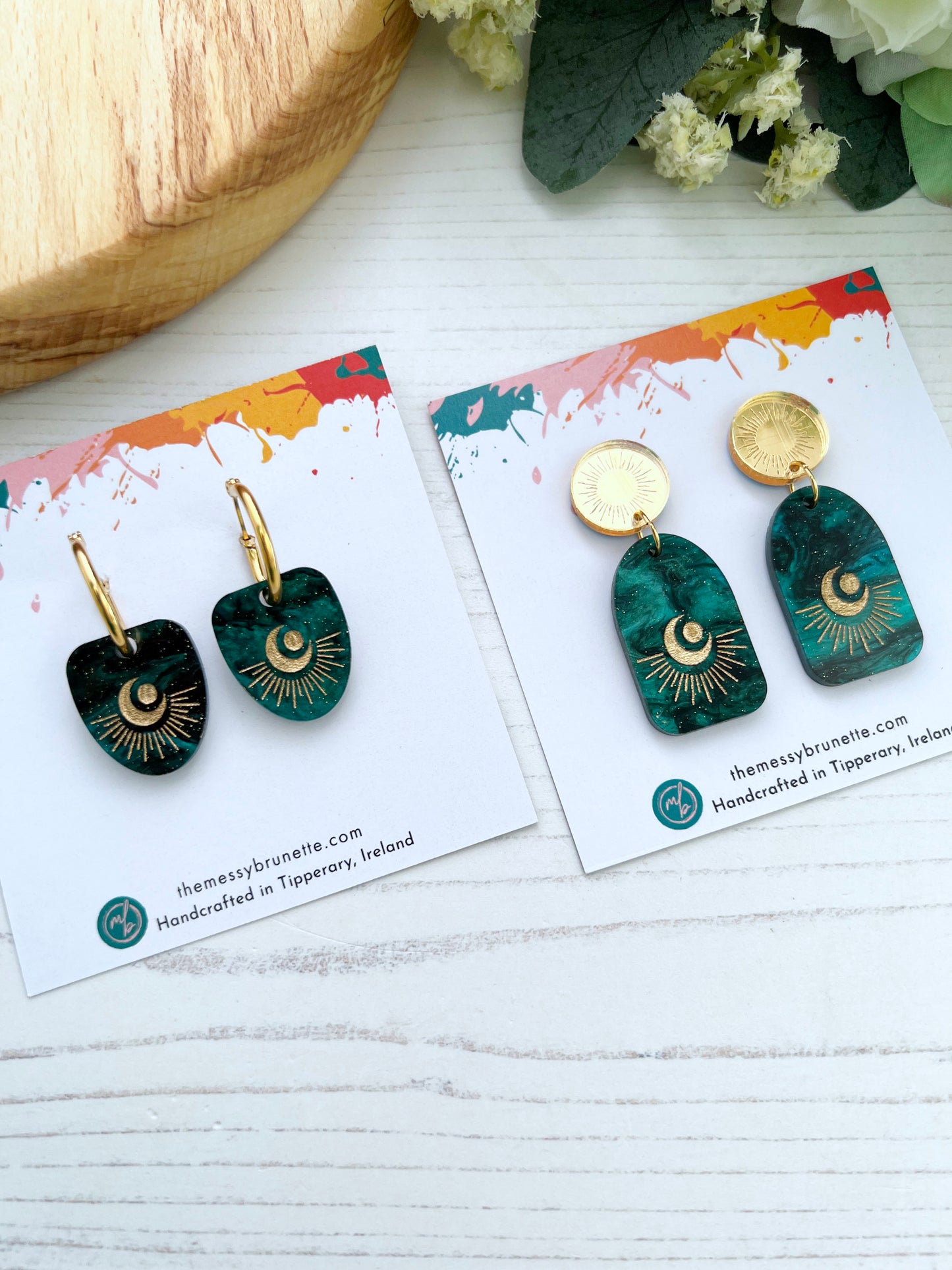 Pair of green and gold celestical earrings with gold accents on a branded card, placed on a white surface with flowers in the background.