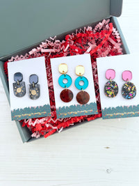 Three pairs of acrylic earrings on cards in a green gift box with red crinkle paper