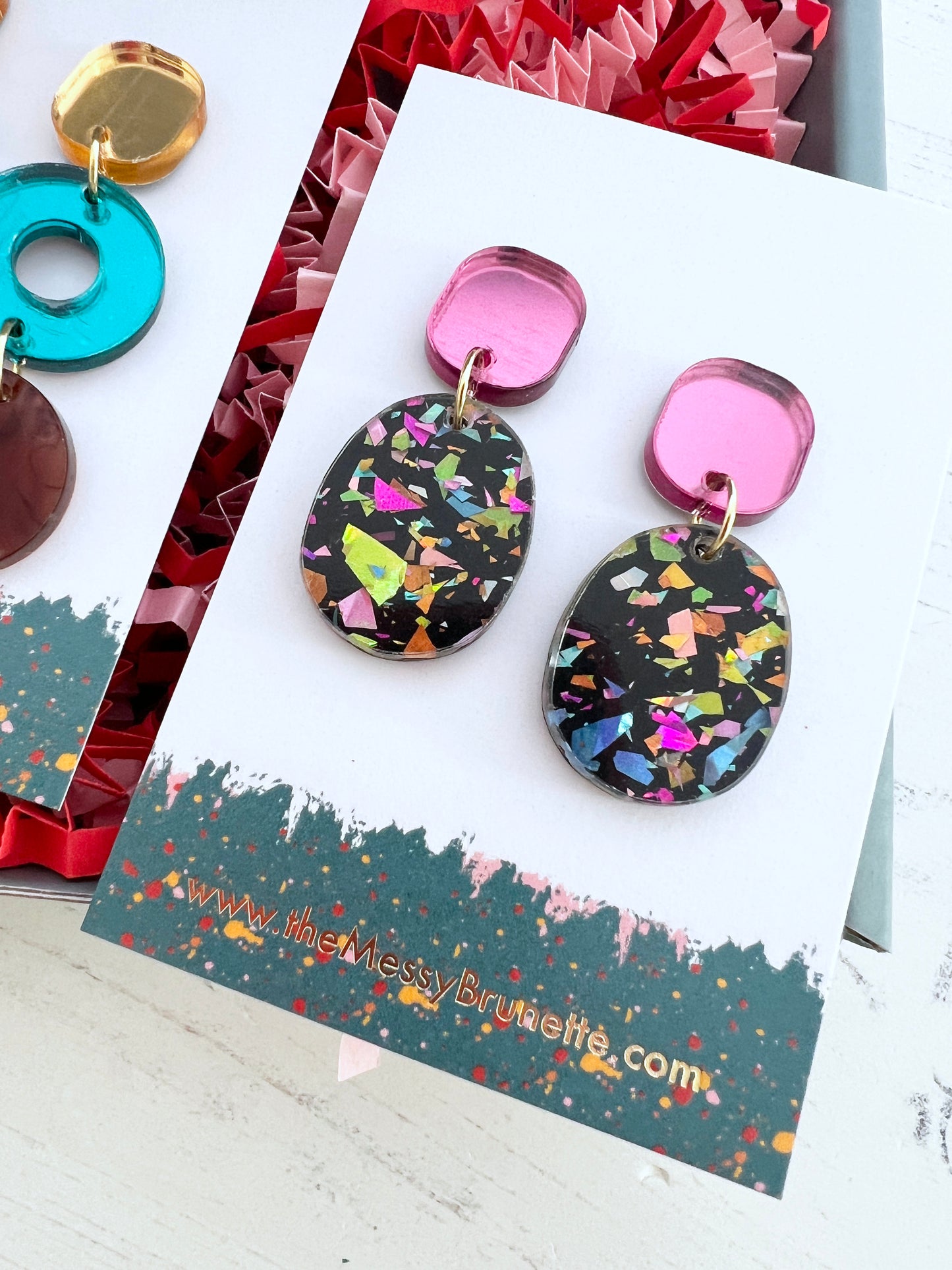 Colourful glitter earring drops on pink studs on a branded card with a colourful background