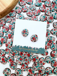 A pair of oval stud Christmas earrings with a red, teal, and white floral pattern, displayed on a white card. The card is surrounded by many matching earrings scattered on a white surface, with a wooden block visible in the top-left corner.