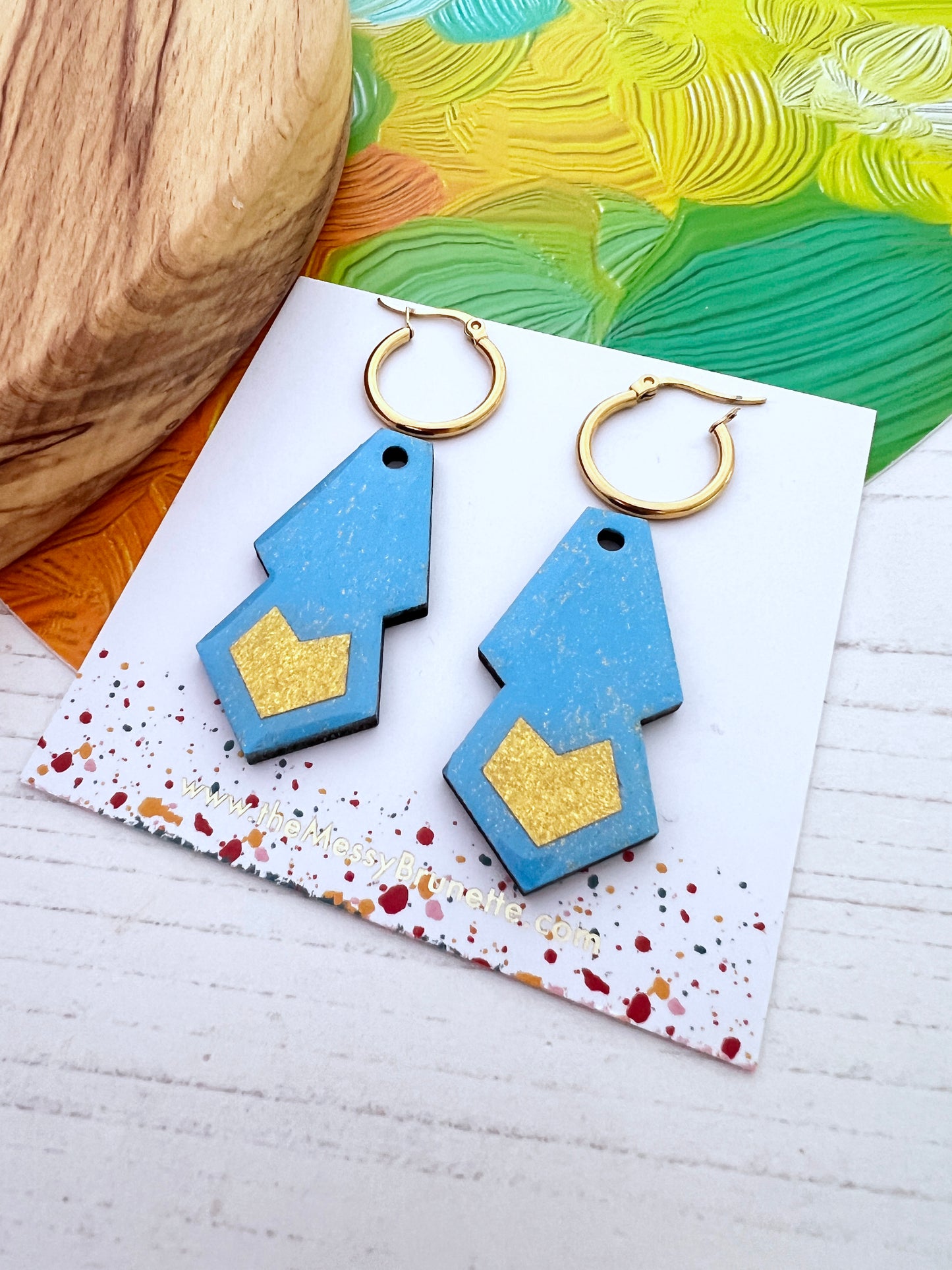 Blue and gold geometric earrings on a colourful card with a wooden surface and green fabric in the background.