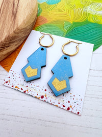 Blue and gold geometric earrings on a colourful card with a wooden surface and green fabric in the background.