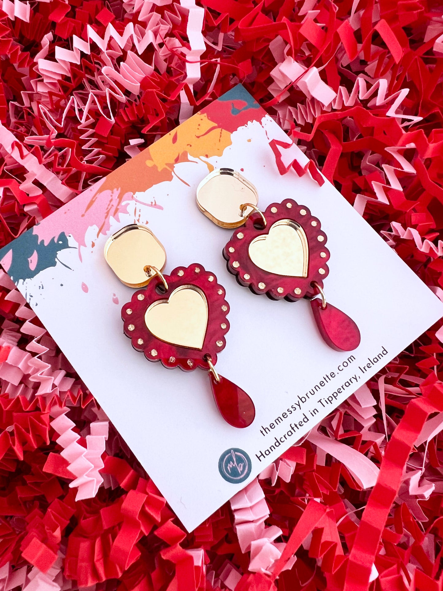 Heart-shaped earrings on a branded card with red and pink crinkled paper background