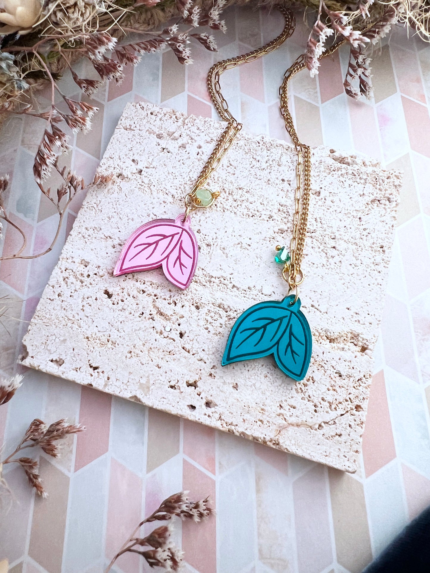 Necklace with pink and blue leaf pendants on a textured surface with dried flowers.