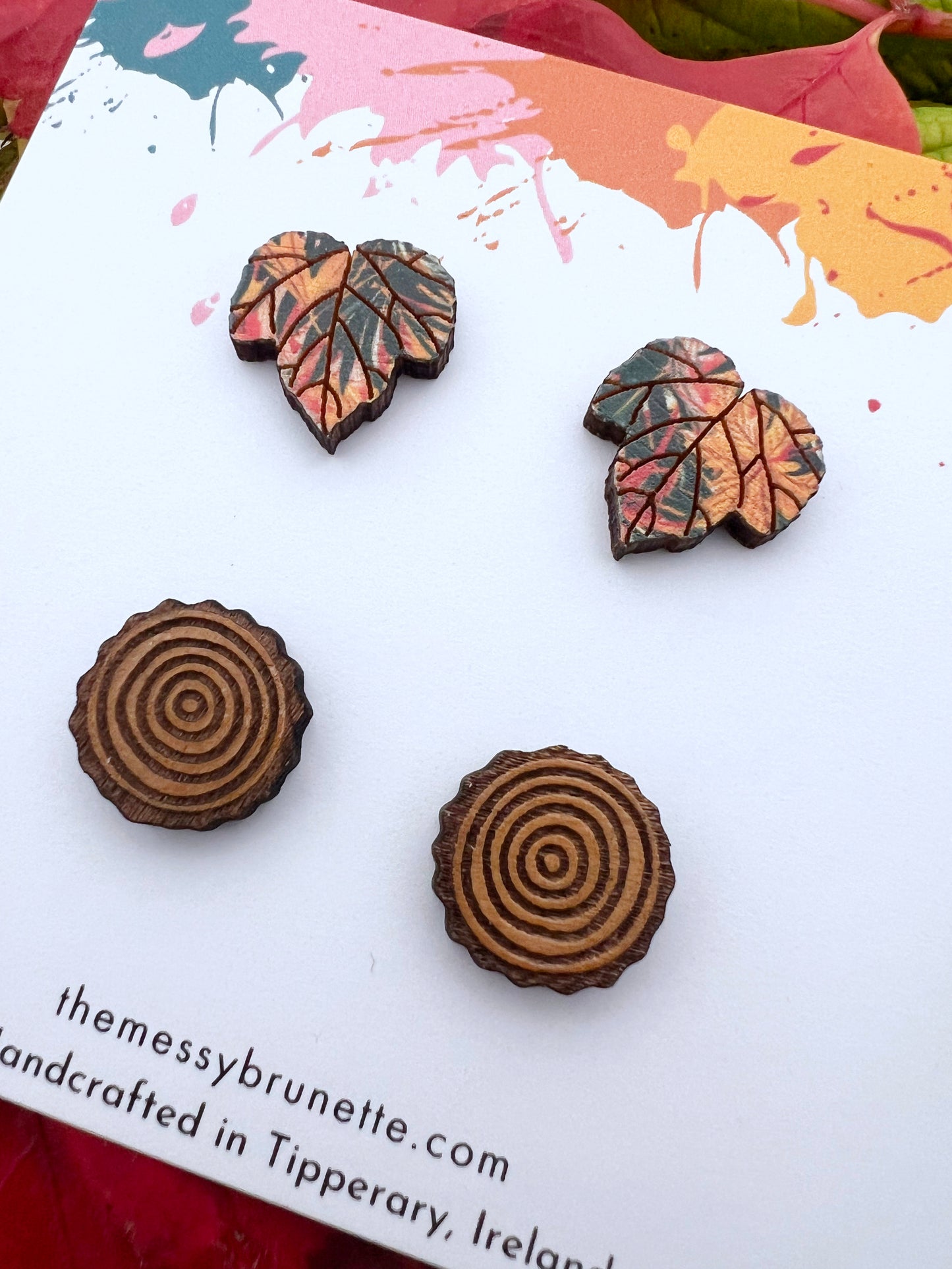 Pair of wood slice studs and maple leaf studs earrings on a card with autumn leaves and acorns in the backgroun