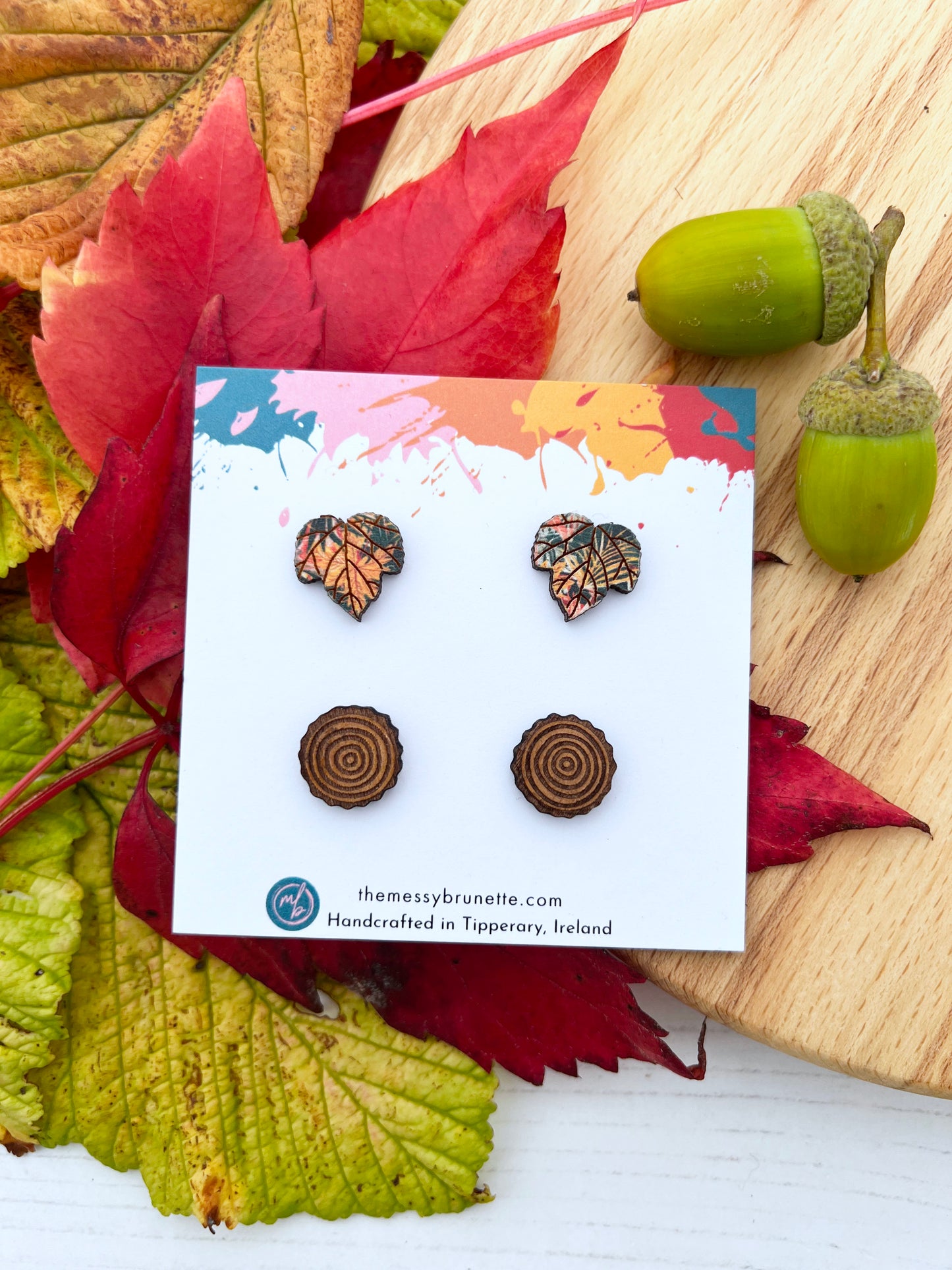 Pair of wood slice studs and maple leaf studs earrings on a card with autumn leaves and acorns in the backgroun