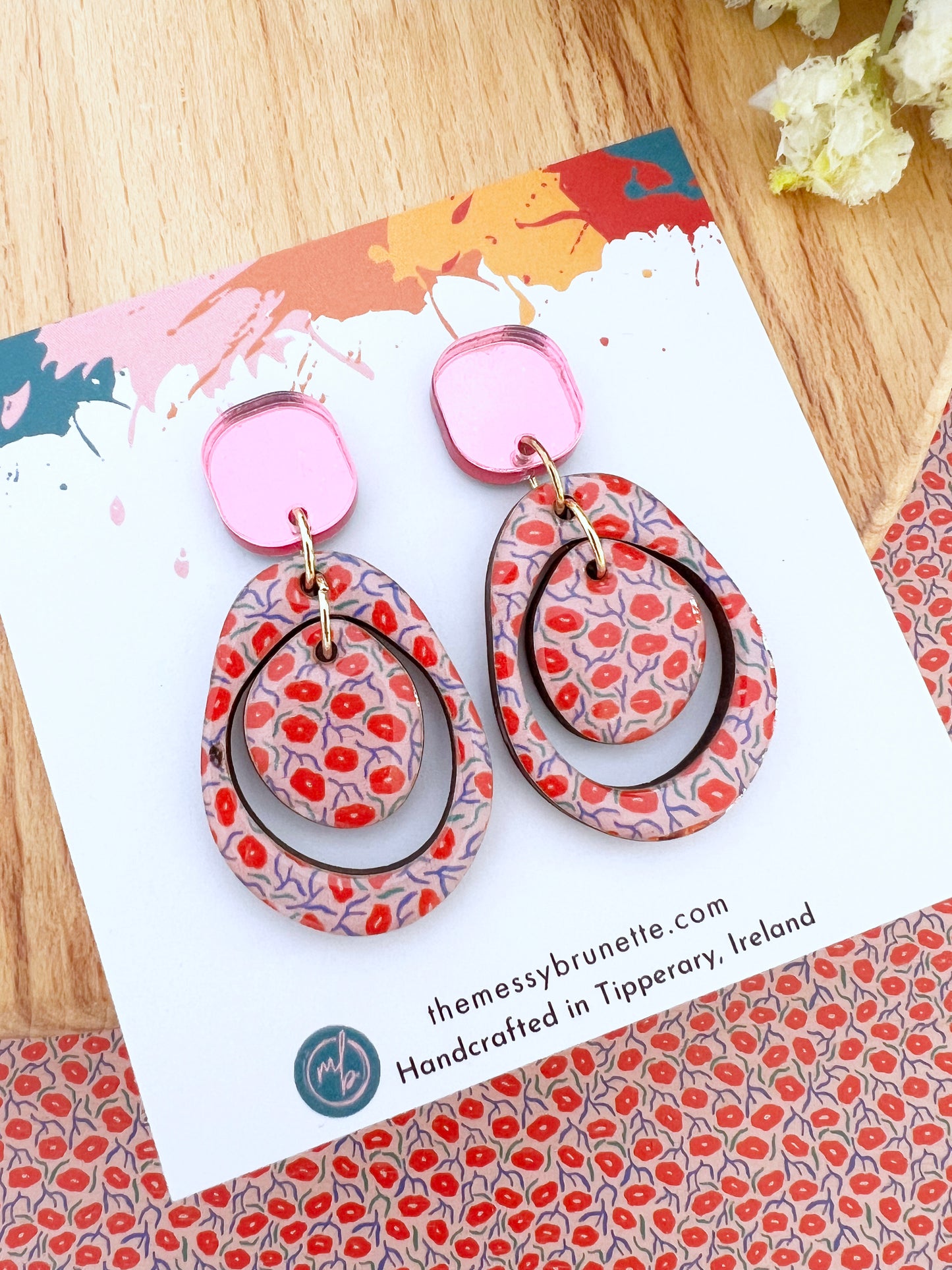 Pair of colorful earrings in red and pinkk on a patterned card with 'themessybrunette' branding.