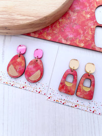 Pair of pink and orange colour clash dangle earrigs on a white surface with a wooden object and pink fabric in the background.