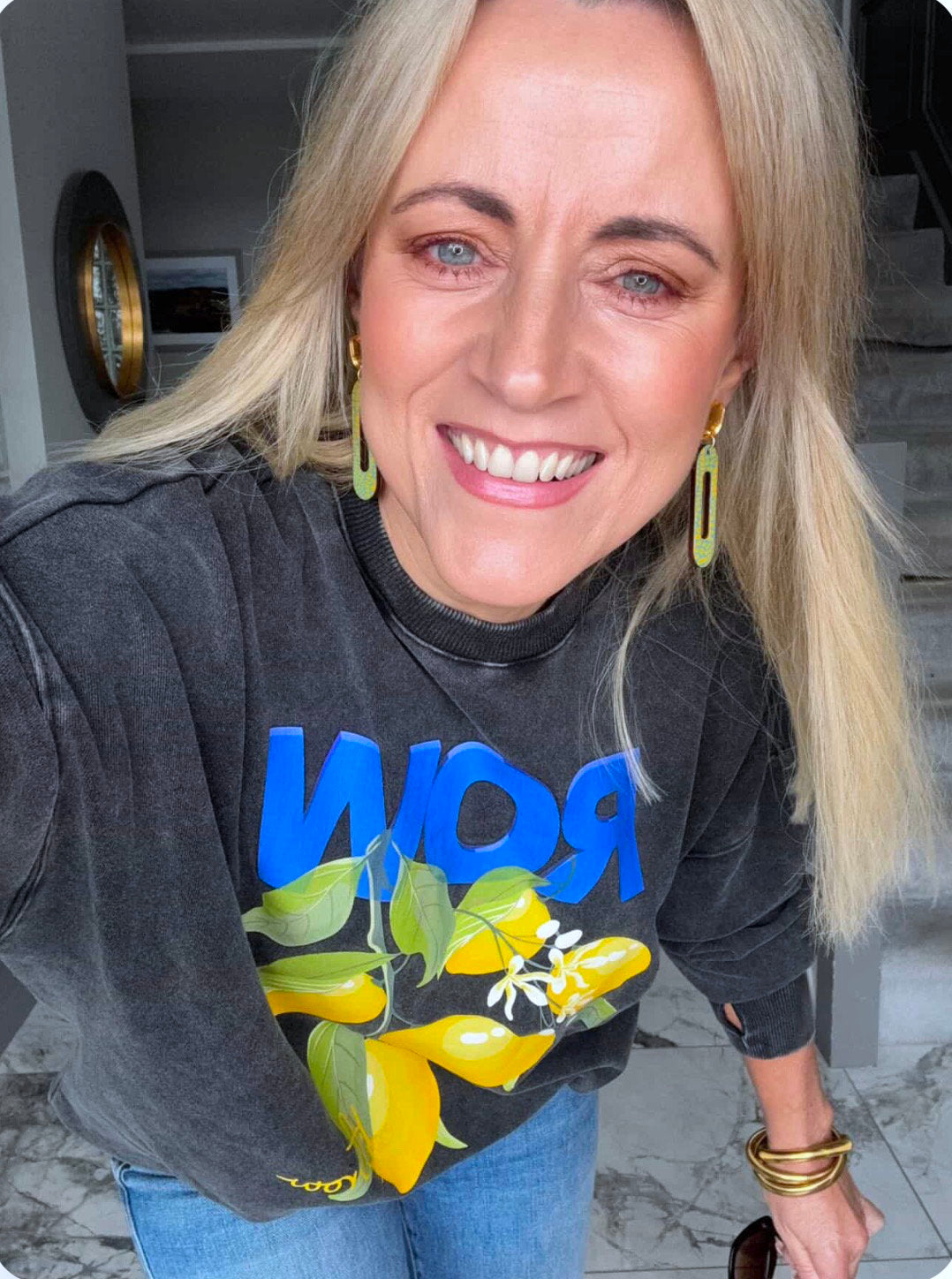 Woman wearing a blue and yellow earrings with a black sweatshirt with colourful design indoors