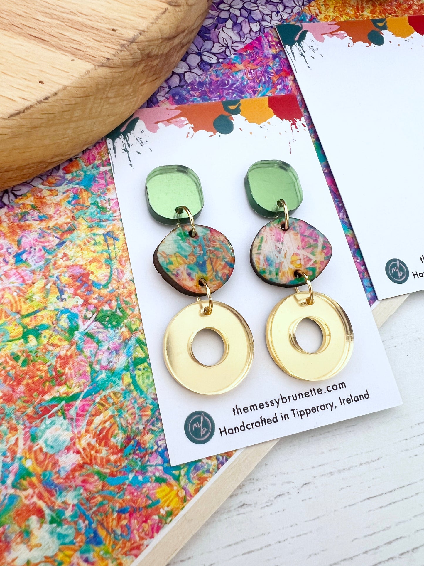 Colourful earrings in green and gold on a branded card with a colourful background
