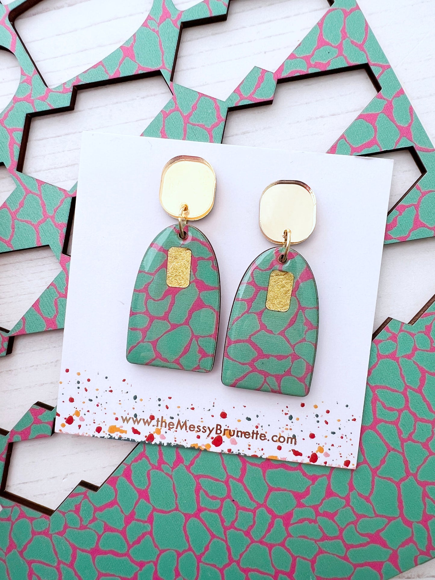 Green and pink patterned earrings on a branded card with geometric shapes in the background.