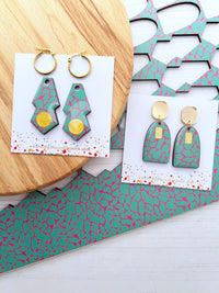 Green and pink patterned earrings on display with a wooden surface and packaging.