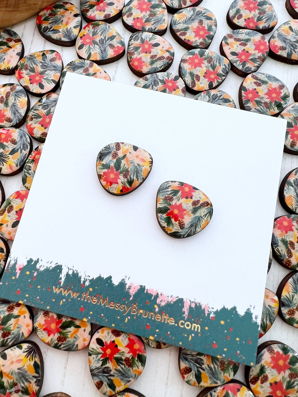 Two wonky shaped Christmas themed stud earrings with a colourful floral and leaf pattern, displayed on a white card with a teal painted border. The card is surrounded by many matching earrings on a white wood surface.