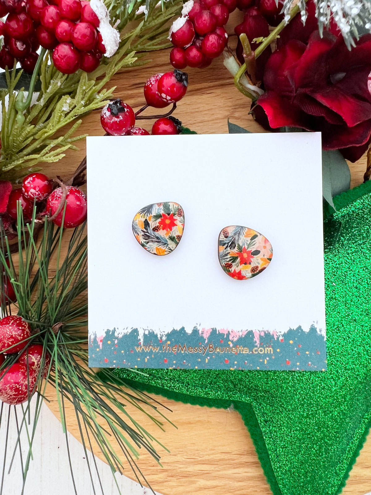 A pair of wooden stud earrings with a colourful Christmas floral pattern, displayed on a white card with a holiday-themed background of pine branches, red berries, and a green glitter star.