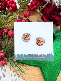 A pair of wooden stud earrings with a colourful Christmas floral pattern, displayed on a white card with a holiday-themed background of pine branches, red berries, and a green glitter star.