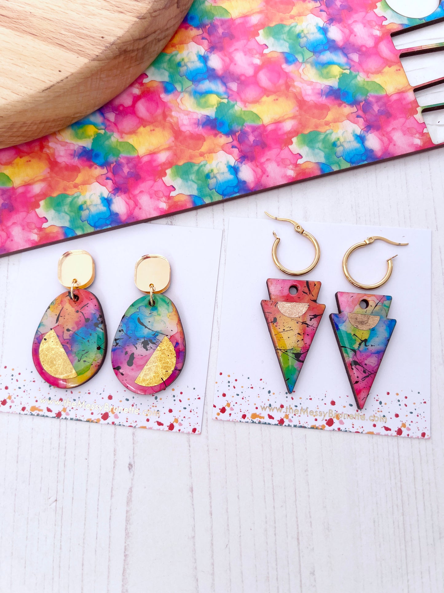 Multi colourful earrings on a white surface with a tie-dye patterned mat in the background.