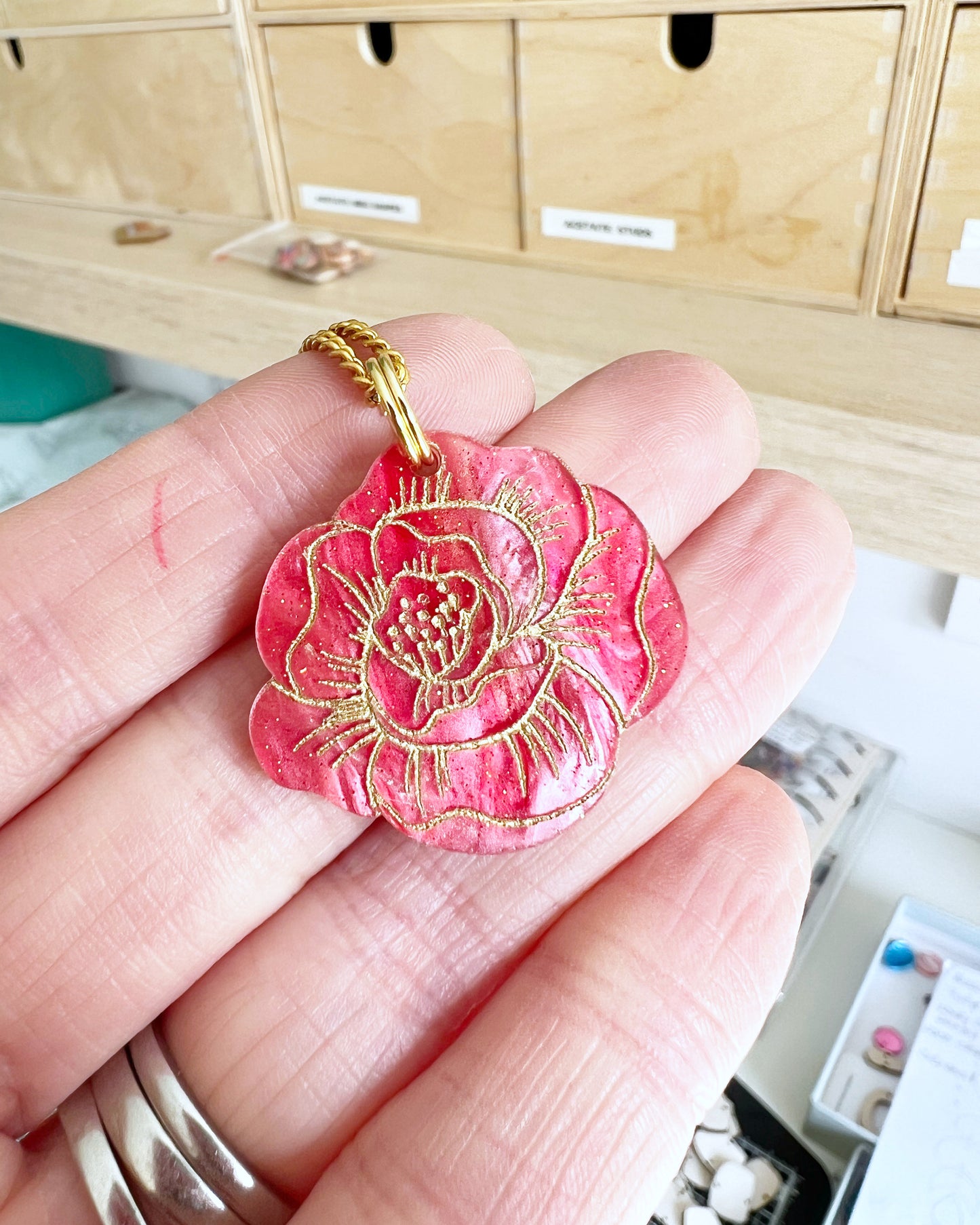 Pink Rose flower-shaped pendant with gold detailing on a hand, with a blurred background.