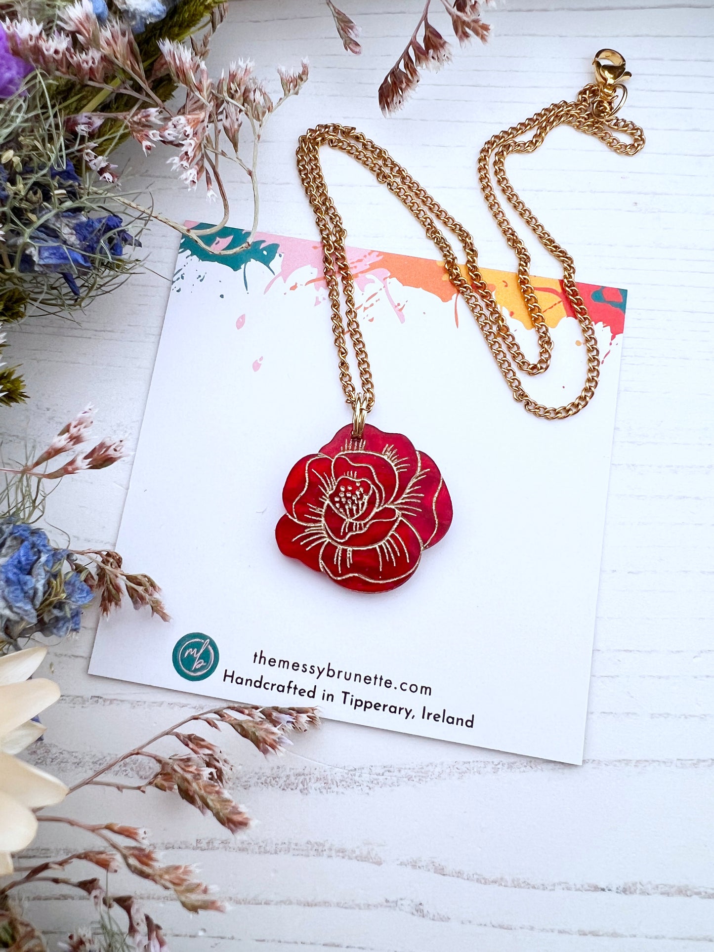 Gold chain necklace with a red rose floral pendant on a white card with branding text, surrounded by dried flowers.