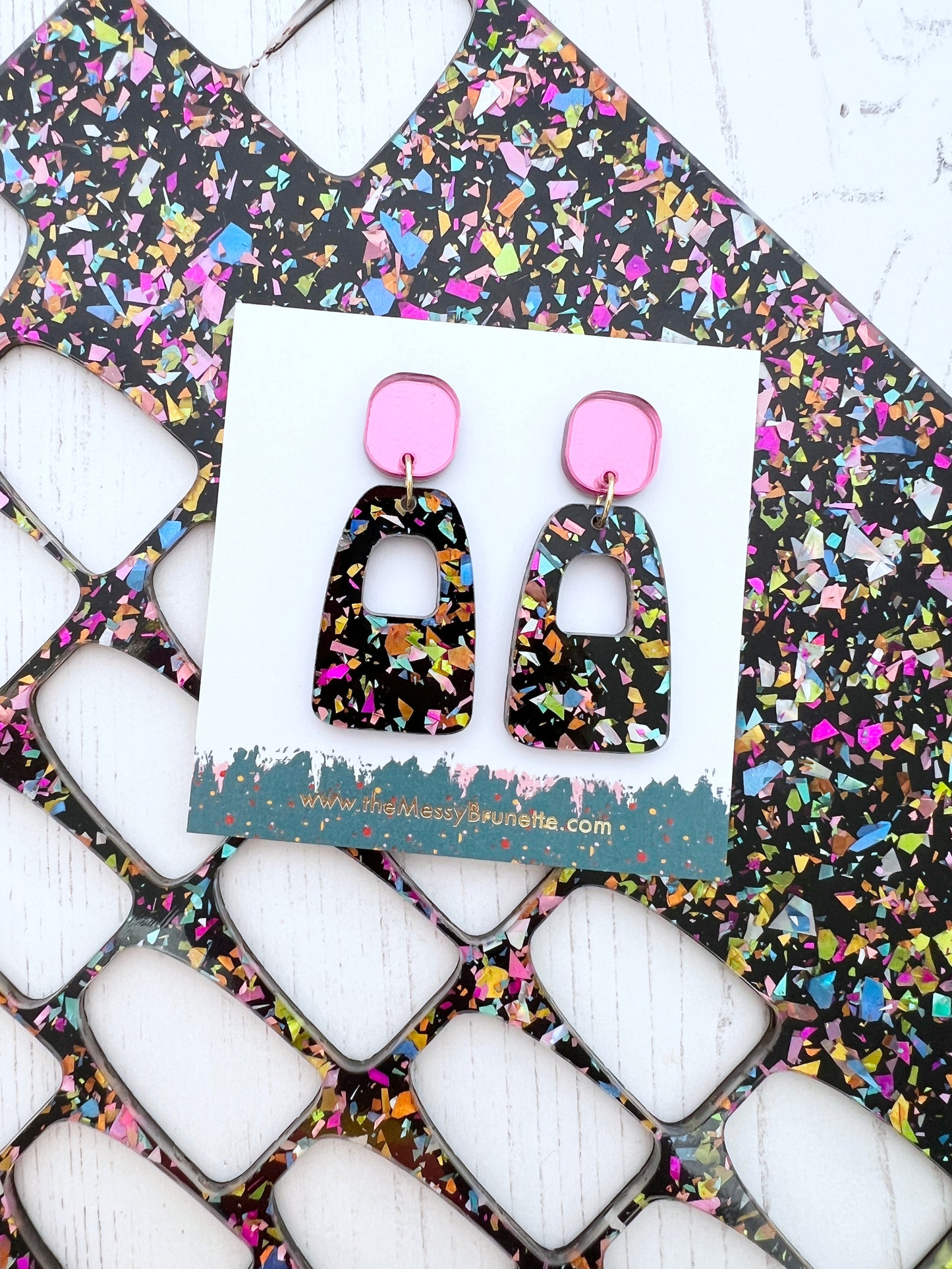 Pair of glitter shards patterned earrings with pink accents on a branded The Messy Brunette card, placed on a colourful abstract background.