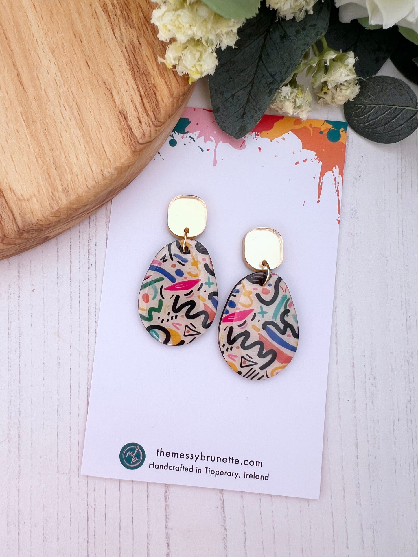 Colourful earrings on a card with 'themessybrunette.com' branding, placed on a wooden surface with flowers.