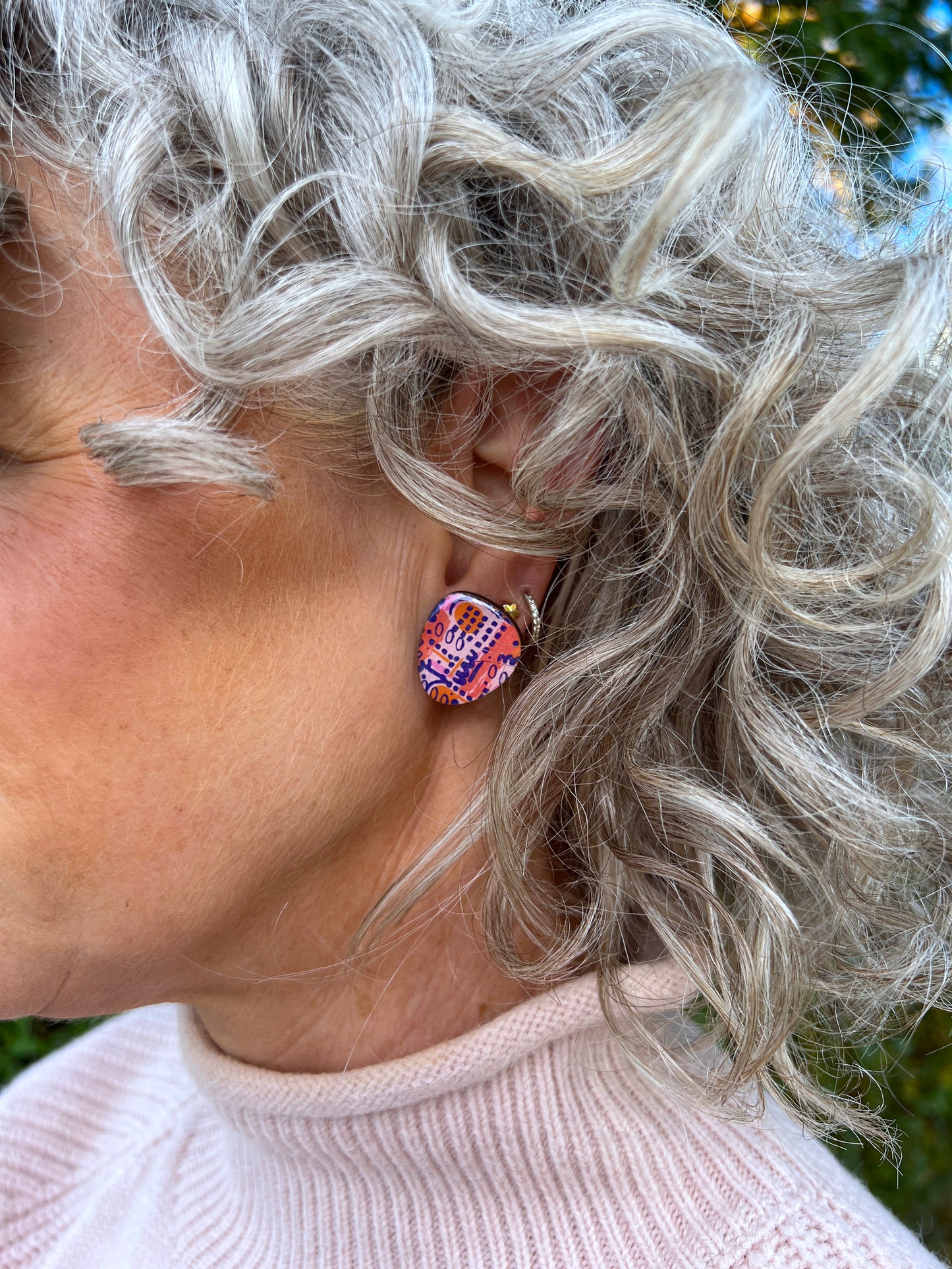 Close-up of a person wearing coluorful statement stud earrings with curly gray hair.