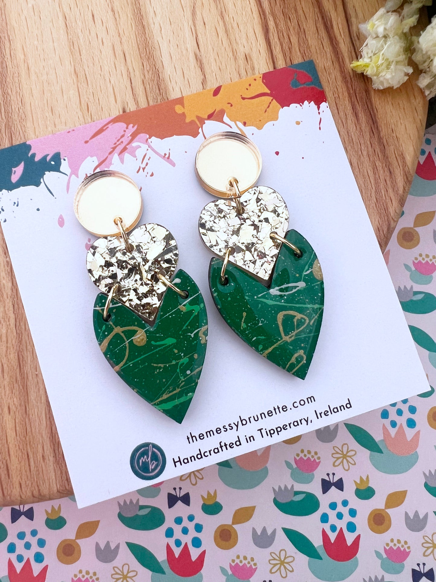 Heart-shaped earrings in green and gold on a branded card with floral background