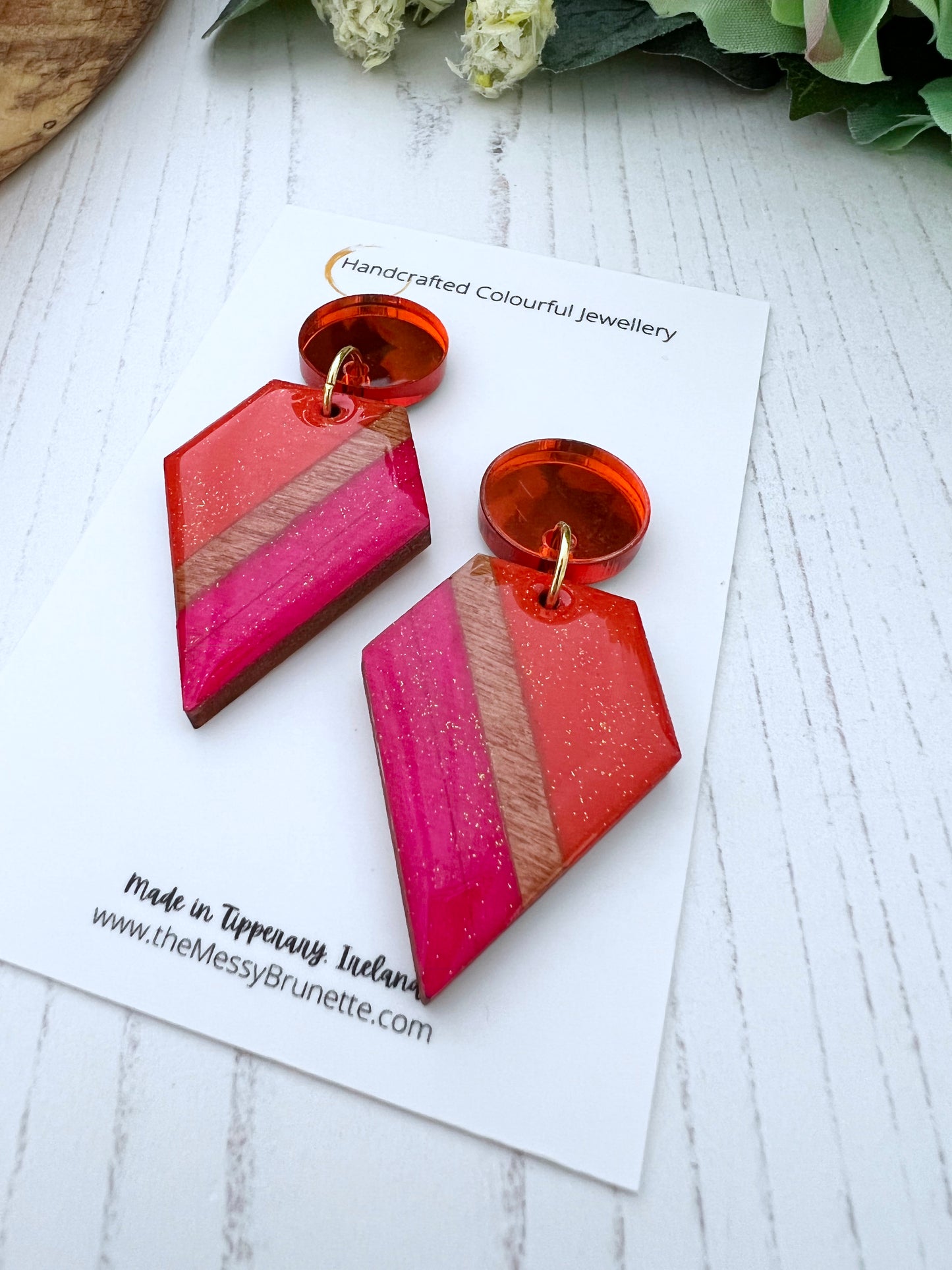 Geometric pink and orange earrings on a white card with branding text.