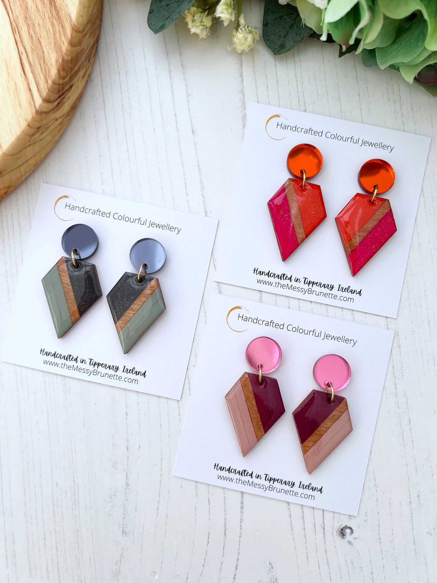 Colourful geometric earrings on display cards with a wooden surface and flowers in the background.