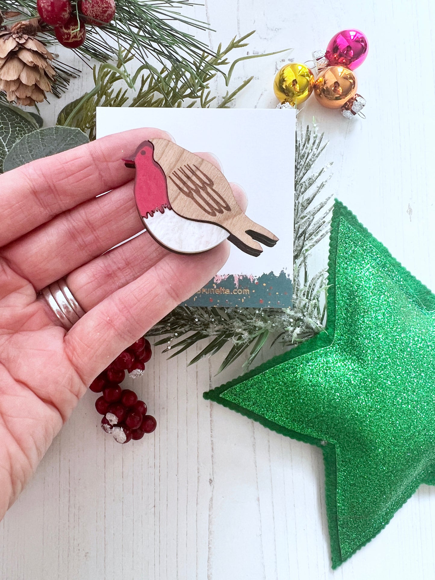 Hand holding a wooden robin brooch with a red body, surrounded by Christmas decorations on a white surface.