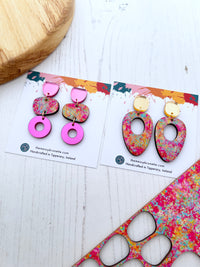 Colourful pink abstract painted wood earrings on a branded card with a wooden block and colourful fabric in the background.