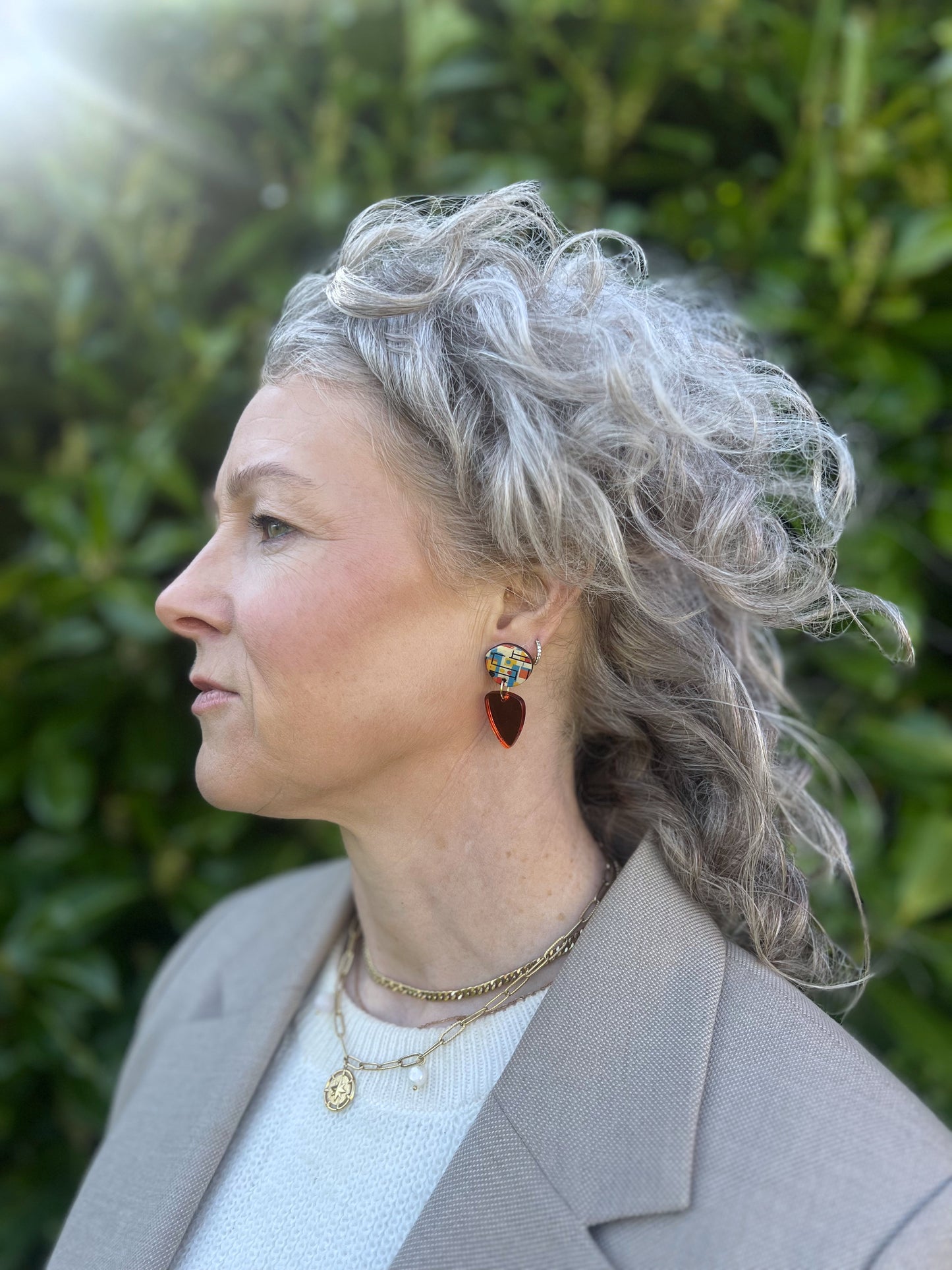 Woman with gray hair wearing earrings and a necklace, standing outdoors with greenery in the background