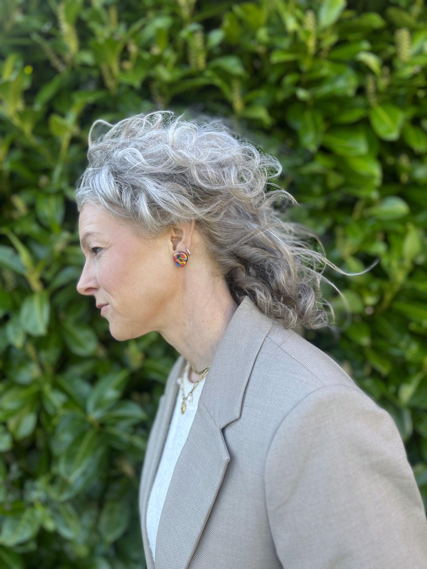 Woman with gray hair wearing large colourful stud earrings a green hedge background