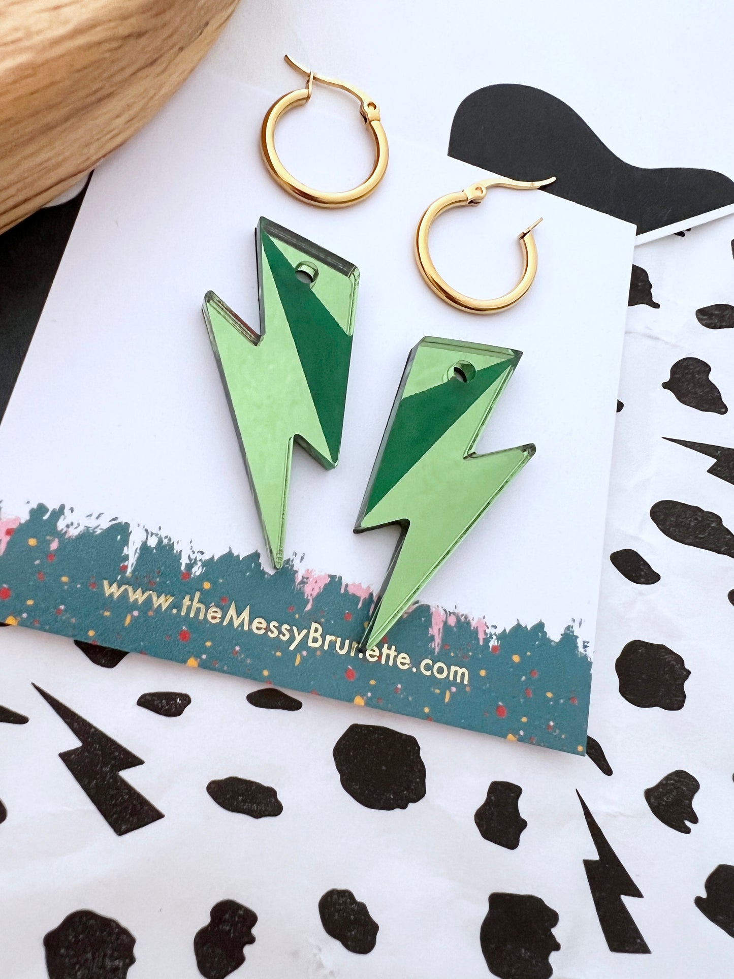 Green lightning bolt earrings on a branded card with gold hoop earrings.