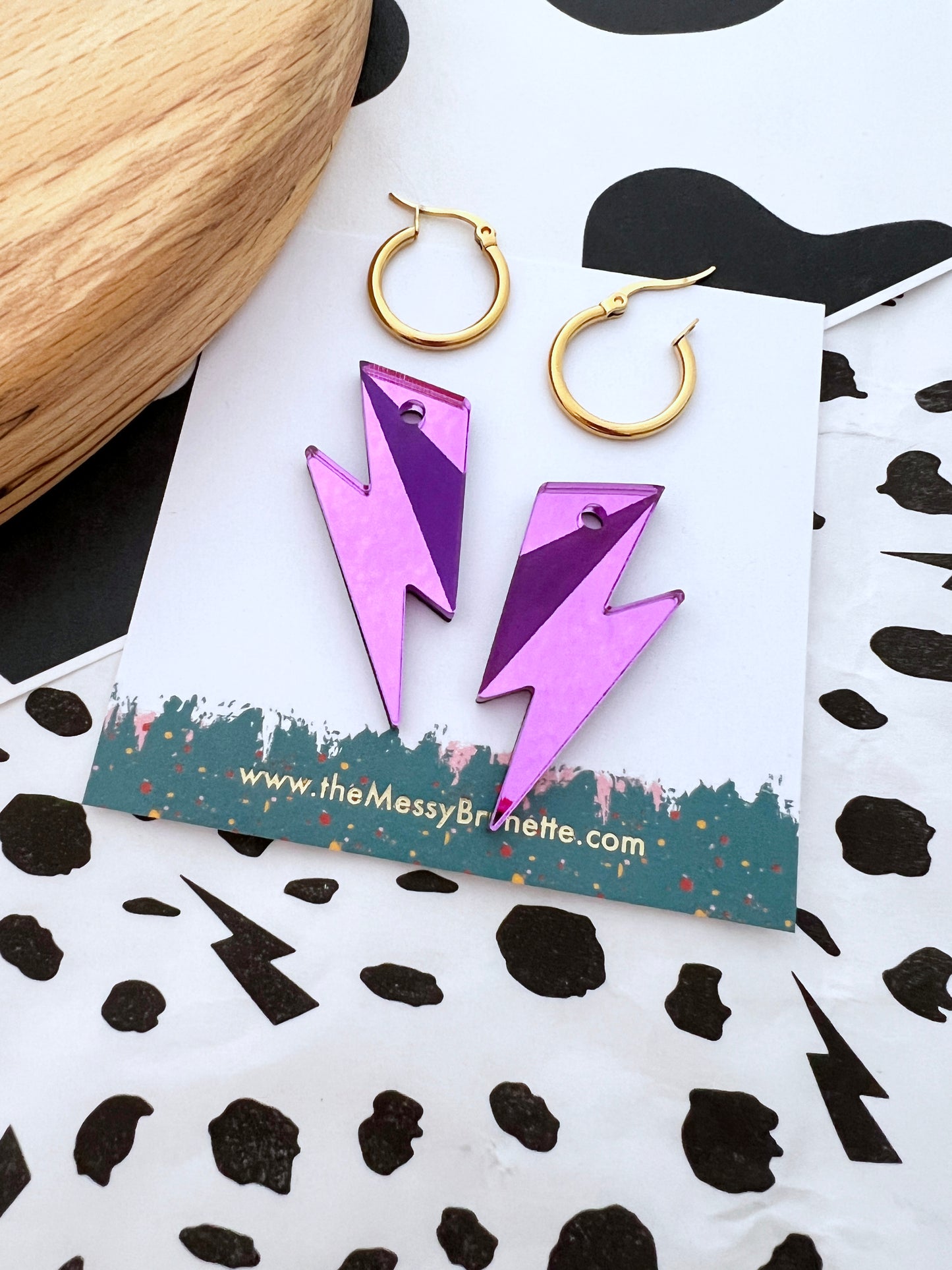 Purple lightning bolt earrings on a branded card with gold hoop earrings and a black and white patterned surface.