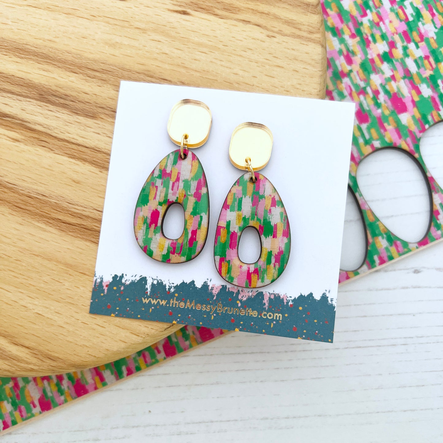 Colourful pink and green earrings on a branded card with a wooden surface and colorful packaging in the background.